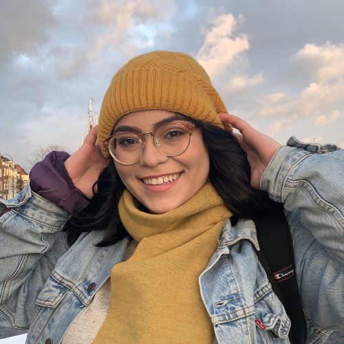 Andrea Argueta smiling wearing glasses, yellow hat, and yellow scarf. 